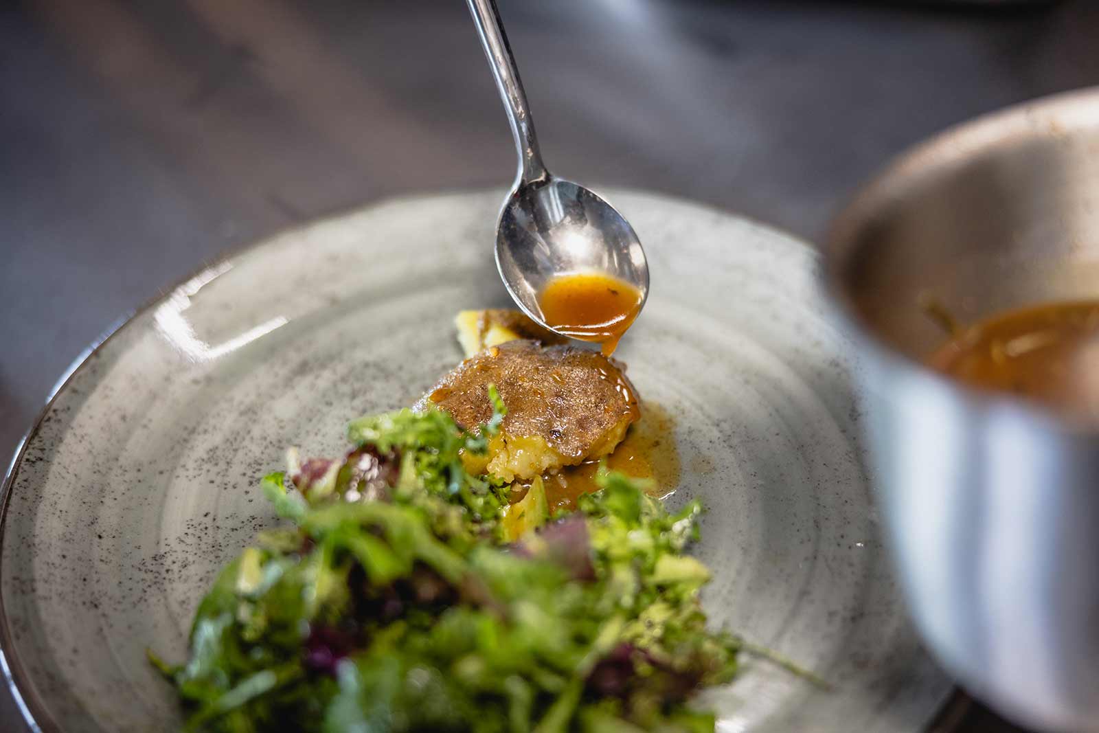 Ein Teller mit einem Stück Fleisch oder Fisch, das mit einer Sauce aus einem Löffel beträufelt wird. Daneben liegt eine Portion grüner Salat. Die Szene befindet sich auf einer grauen Platte.