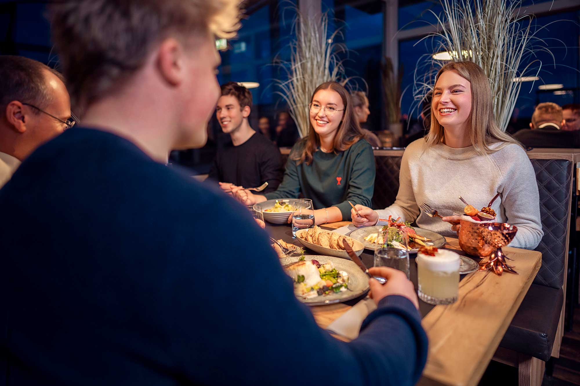 Eine Gruppe von Menschen sitzt um einen Tisch und isst gemeinsam. Auf dem Tisch stehen verschiedene Speisen. Alle scheinen sich angenehm zu unterhalten und zu lachen. Der Raum ist modern eingerichtet.