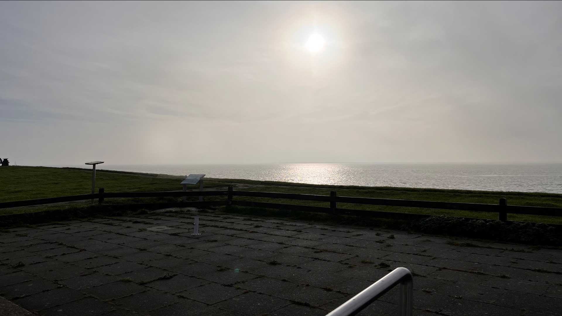 Eine gepflasterte Terrasse mit niedriger Umzäunung blickt auf das Meer. Die Sonne scheint durch einen bewölkten Himmel und spiegelt sich im Wasser. Grünflächen befinden sich links im Bild.