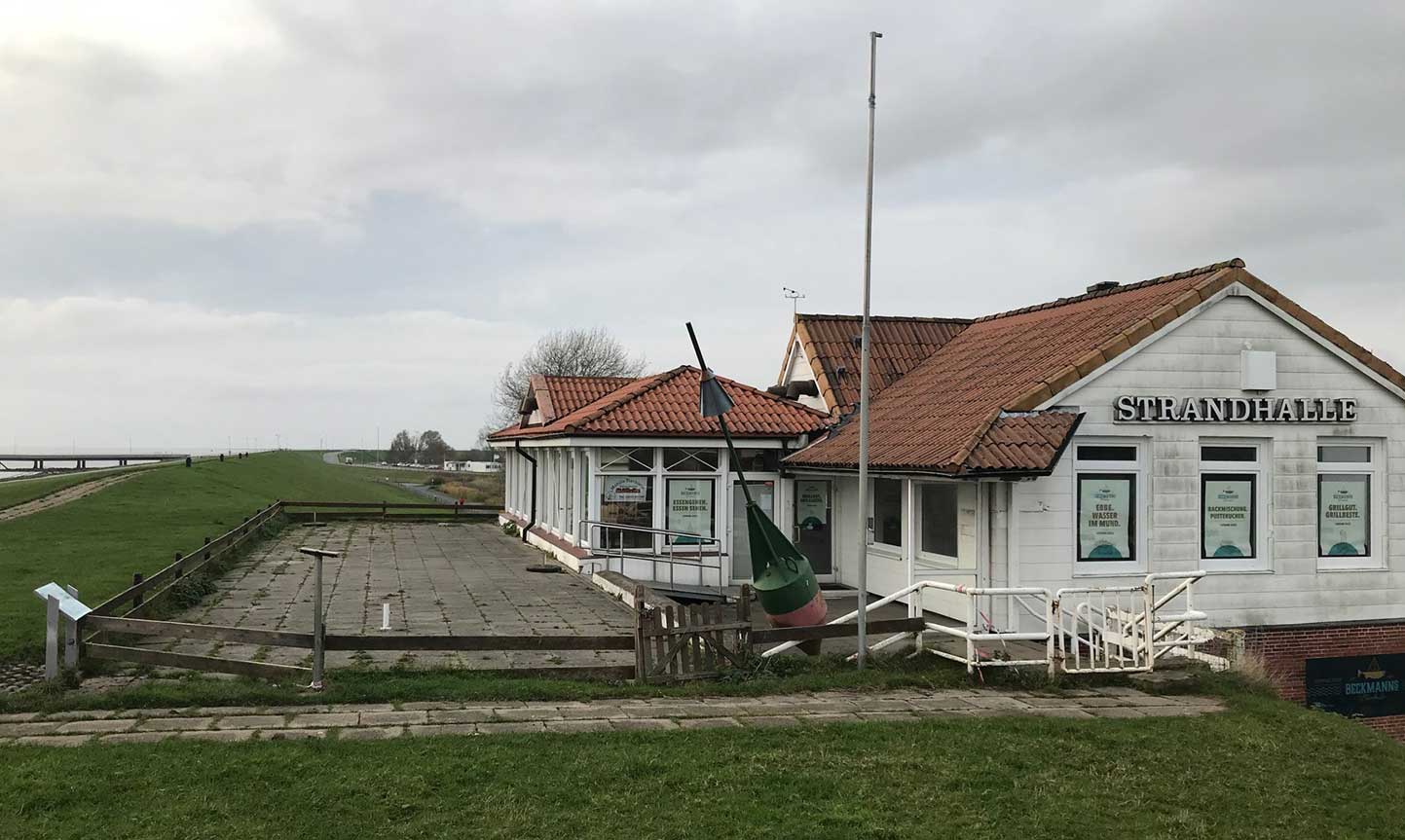 Ein Gebäude mit dem Schriftzug "Strandhalle" steht neben einem gepflasterten Platz mit Holzgeländer. Im Hintergrund ist ein Deich und eine Straße zu sehen. Der Himmel ist bewölkt.