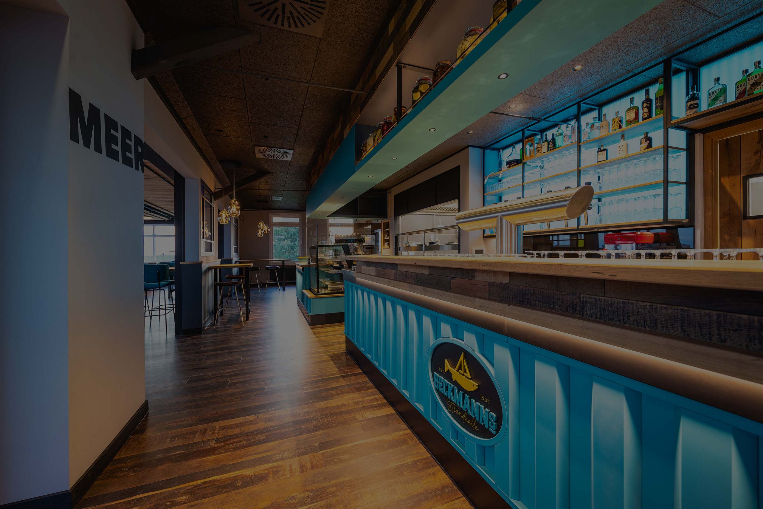 Moderne Bar mit türkisfarbener Front. An der Decke sind dunkle Paneele, der Boden ist aus Holz. Im Hintergrund befinden sich Tische und Stühle. Flaschen sind in Regalen hinter der Bar aufgestellt. Beleuchtung sorgt für ein warmes Ambiente.
