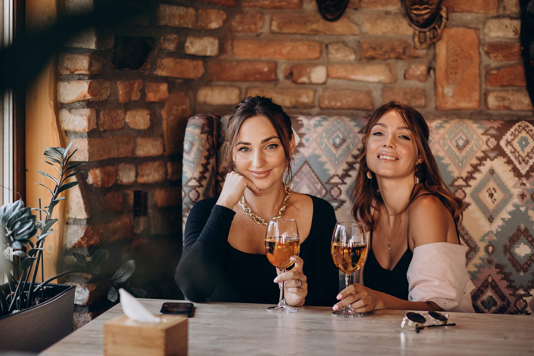 Zwei Personen sitzen lächelnd an einem Tisch vor einer Backsteinwand in einem gemütlichen Restaurant. Sie halten Gläser mit Getränken. Pflanzen und dekorative Elemente sind im Hintergrund zu sehen.